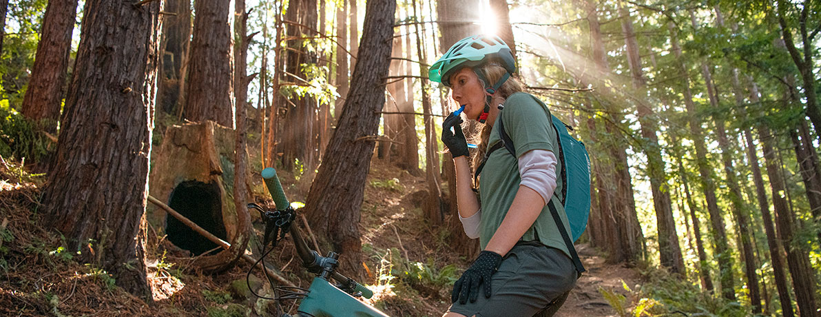 Woman biking in the woods and drinking from a hydration pack.